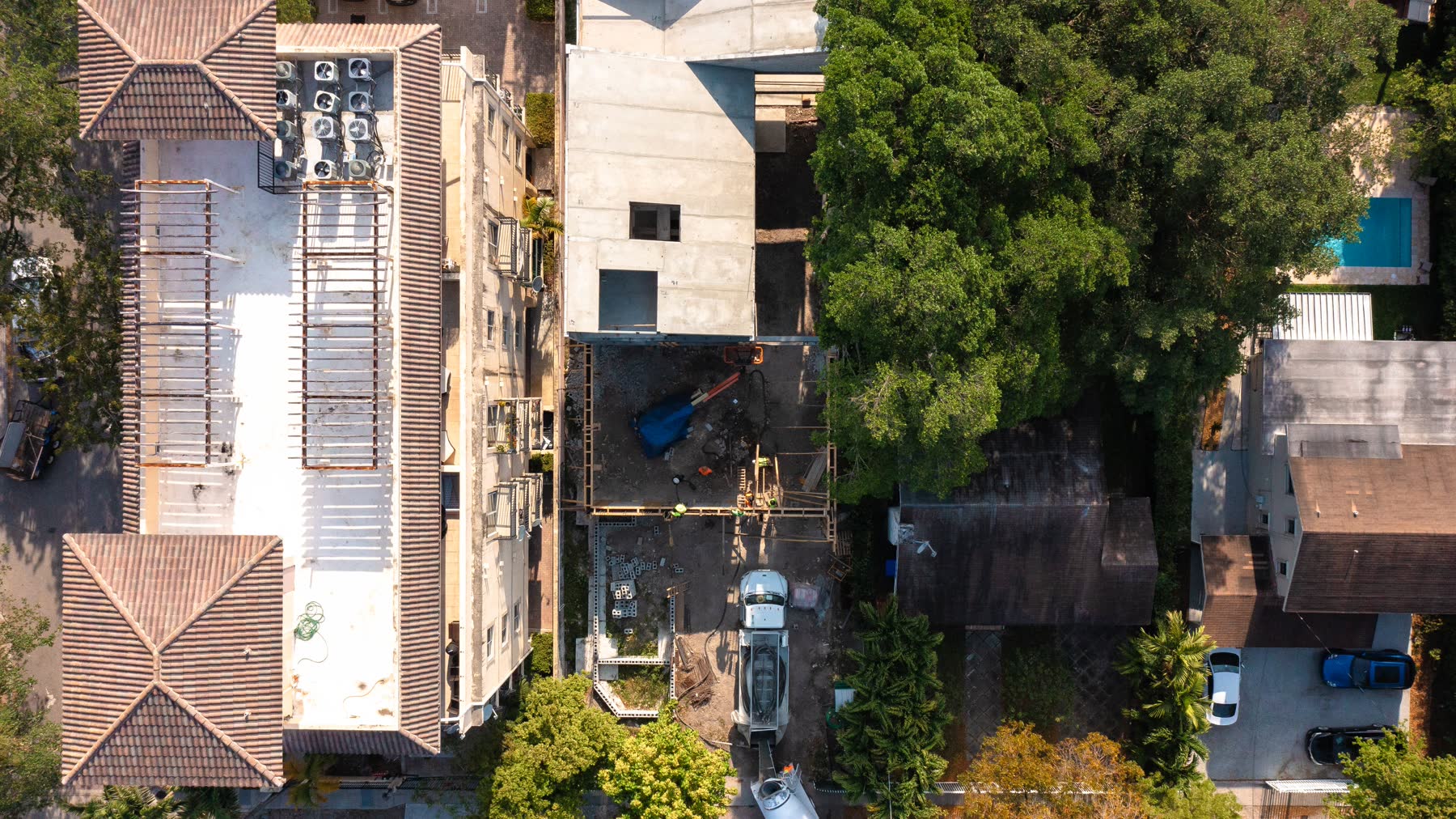 Vista aérea directamente sobre el sitio con la estructura de concreto vaciada en pie, techos vecinos pegados contra ambas colindancias y follaje cerrándose sobre el bloque.