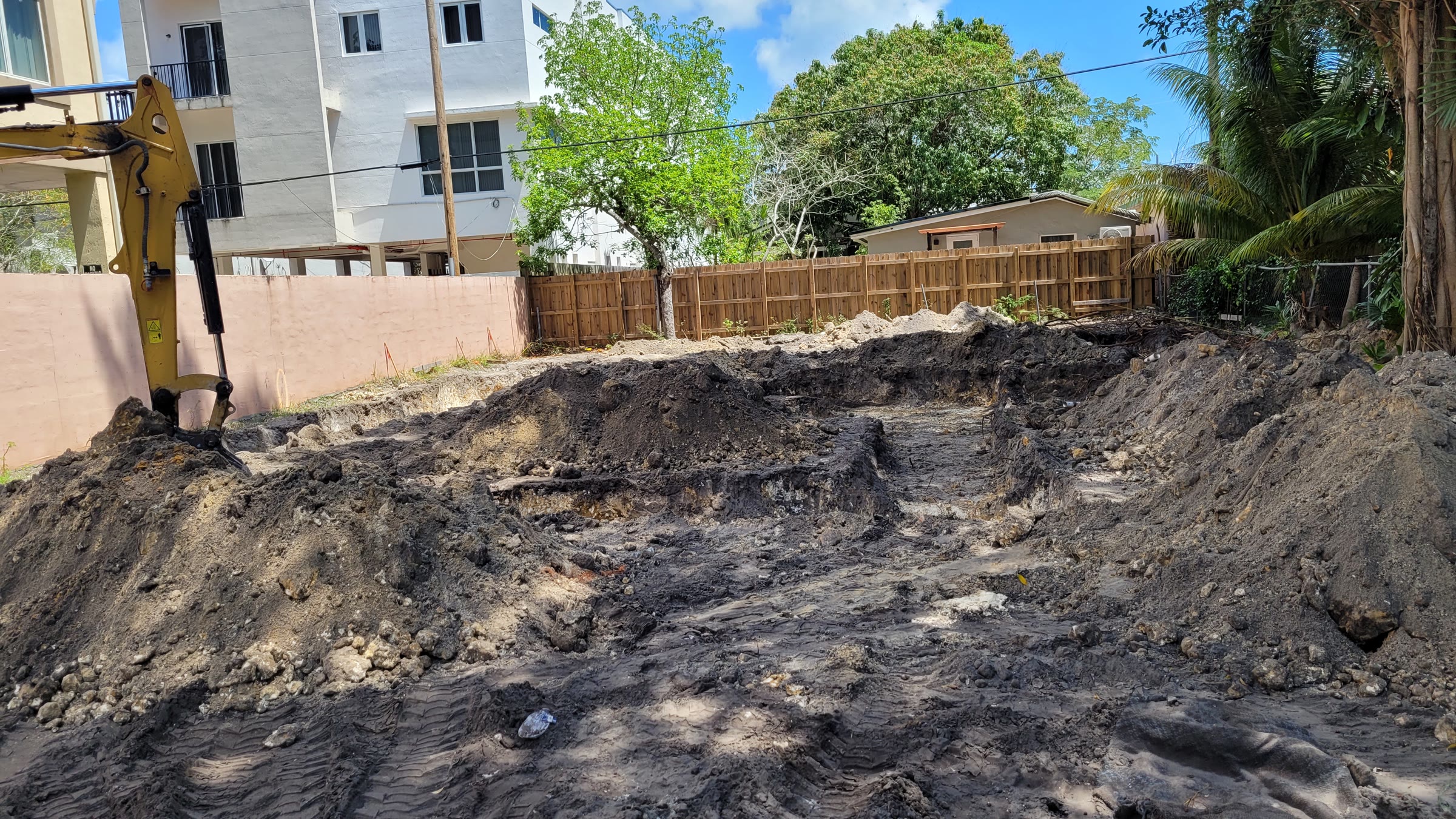 Un brazo de excavadora sobre el lote despejado, con zanjas de zapata esbozadas en relleno oscuro entre las paredes de colindancia, con edificios vecinos cerca a ambos lados.