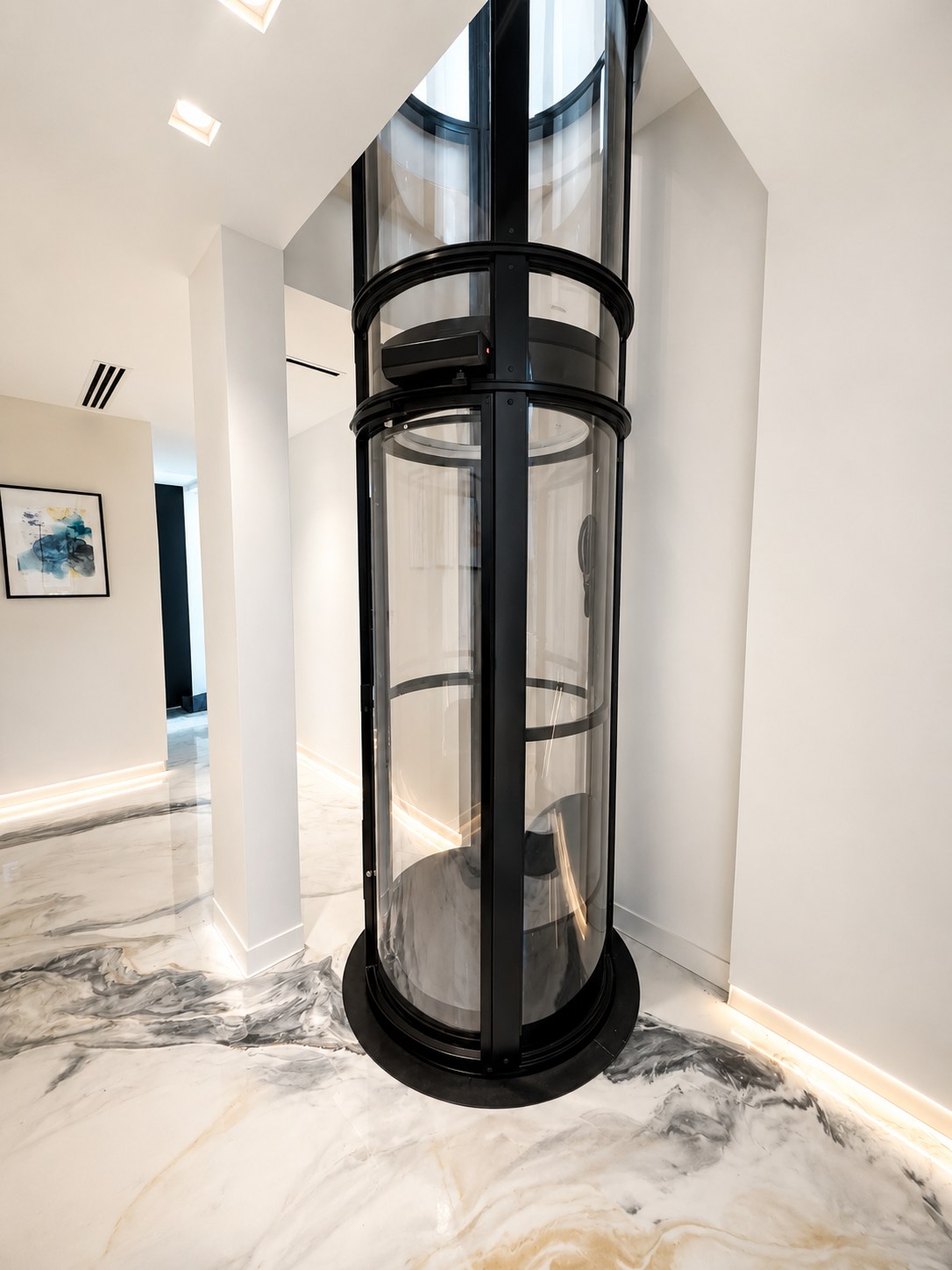 Un elevador neumático de vacío cilíndrico, de vidrio con marco negro, de piso a techo en un pasillo con piso de mármol.