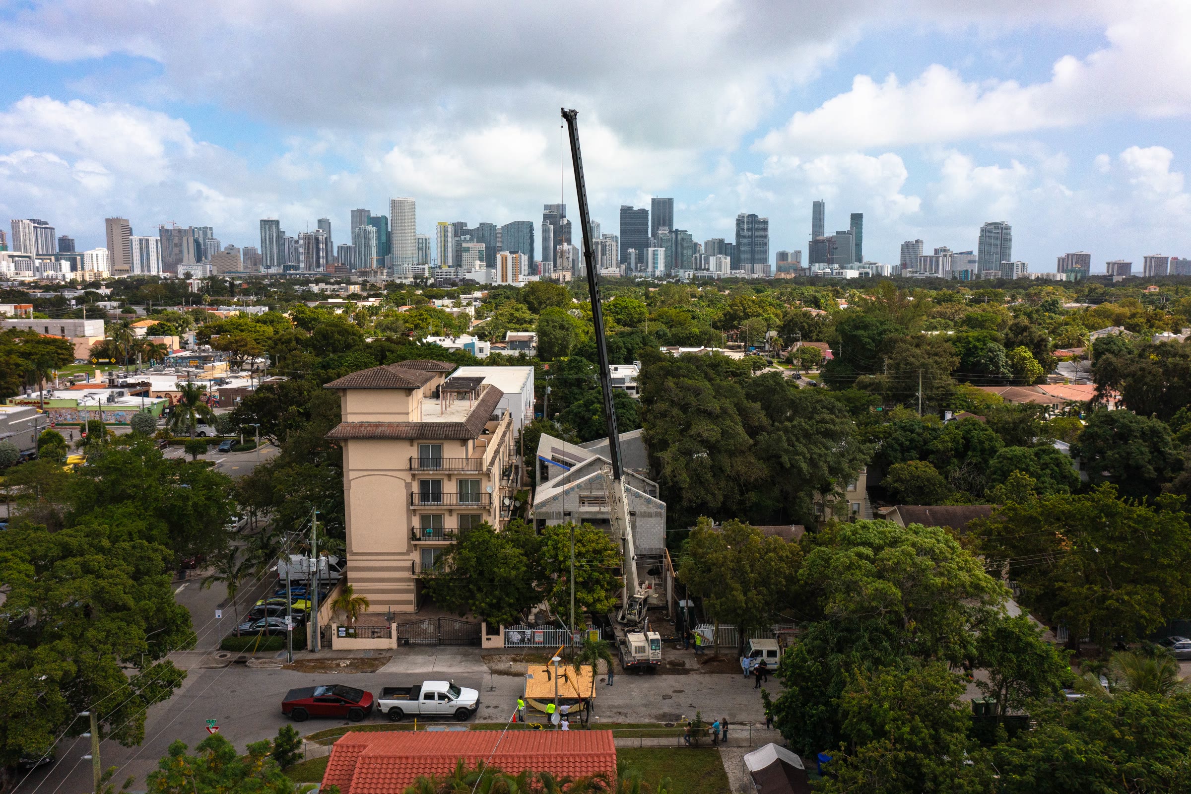 Aerial over the site at shell stage: a crawler crane's boom rising beside the raw concrete and block structure, the neighbouring apartment block hard against it, downtown Miami on the horizon.