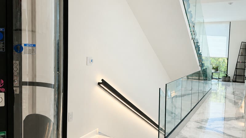 A continuous lit handrail running down a smooth plaster wall beside a frameless glass guardrail.