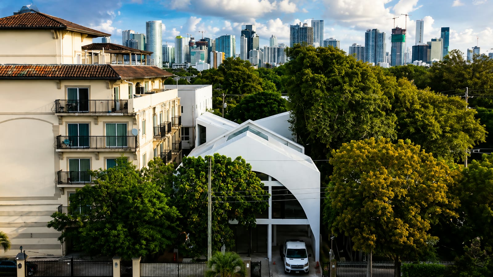 Leslie House en su calle de Little Havana: planos de techo plegados sobre una gran apertura arqueada, un bloque de apartamentos vecino al lado y el horizonte del centro de Miami al fondo.